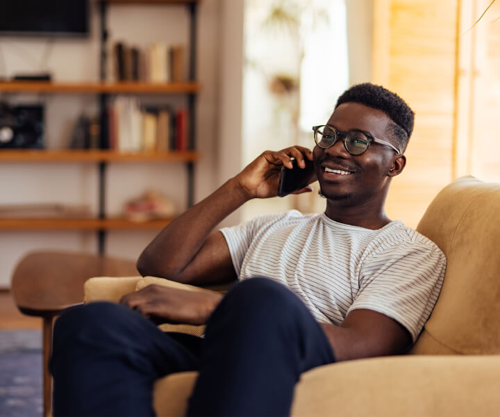 A man sitting on a chair, on his phone smiling. He appears to be in a living room, with bookshelves out of focus behind him.)