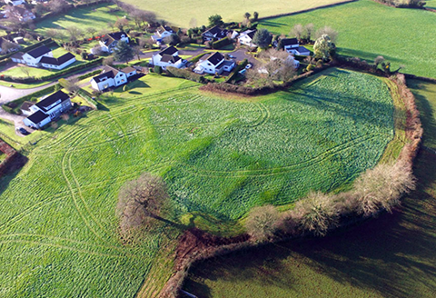 A village hamlet seen from the air. Houses clustered next to lush green fields can be see in the areal shot.)