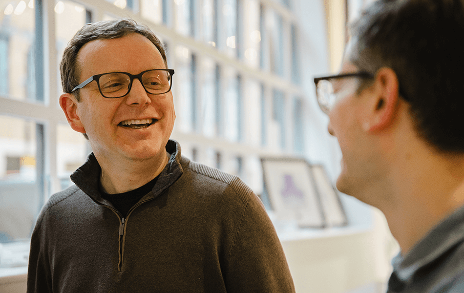 A man in glasses looking away from the camera smiling at another man. The second man is out of focus.)